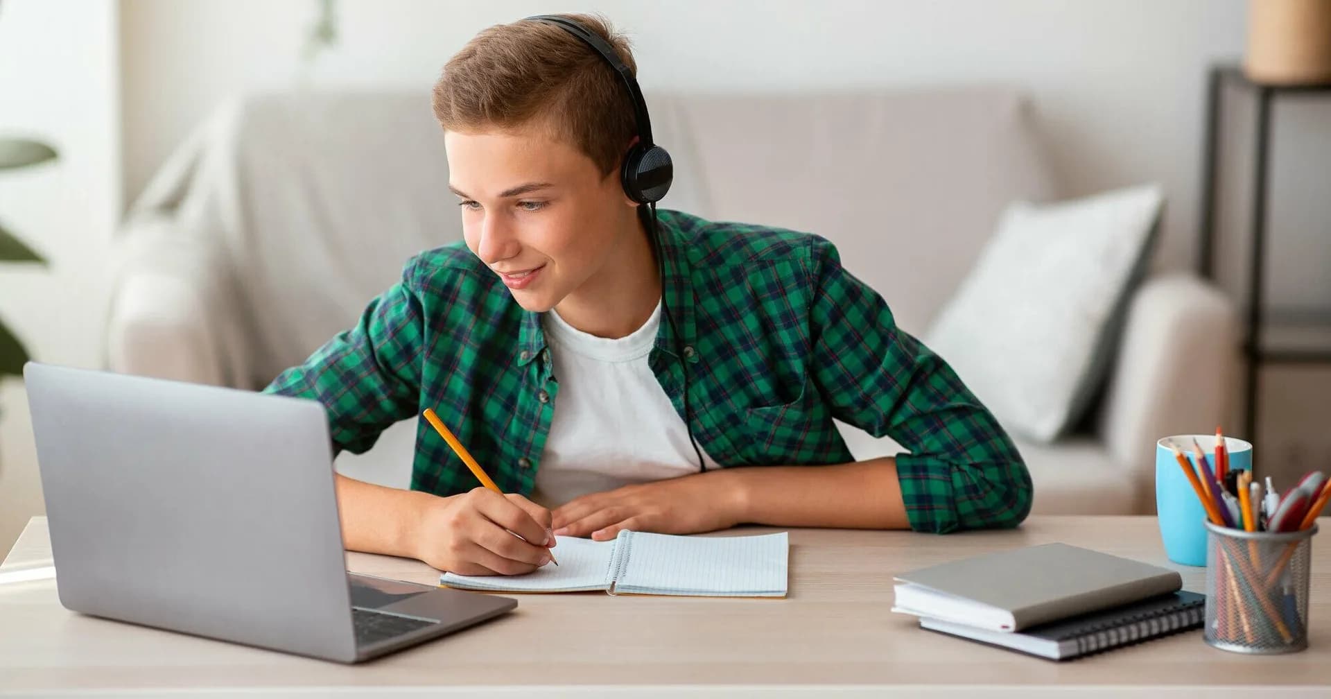 A student wearing headphones takes notes while working on a laptop at home