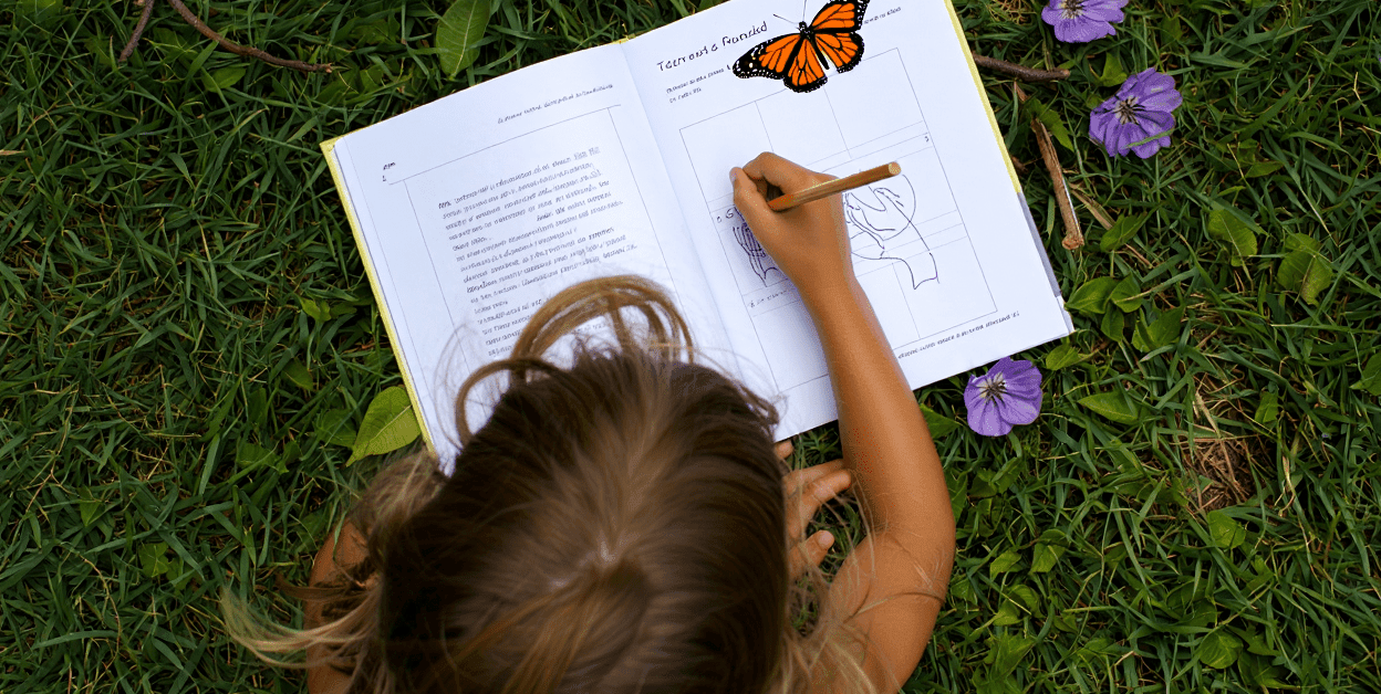 Child drawing in a book on green grass with purple flowers nearby.
