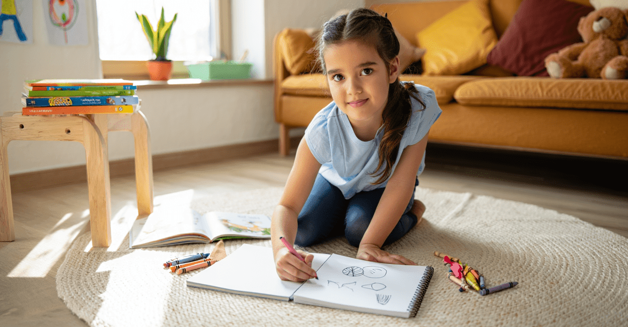 Child drawing in a sketchbook on the floor with crayons nearby.