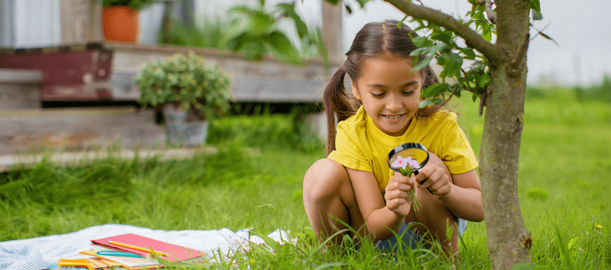 Child in yellow shirt examining pink flowers with a magnifying glass.