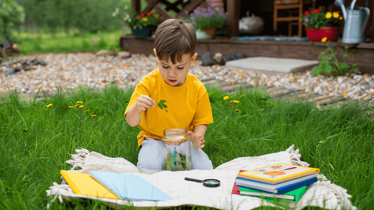 Child in yellow shirt putting a leaf into a jar on a blanket in the grass.