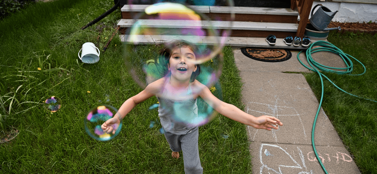 A child joyfully runs through grass, surrounded by colorful bubbles.