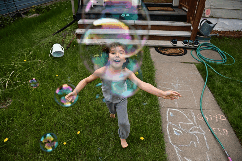 Child running barefoot on grass, surrounded by bubbles, with a sidewalk nearby.