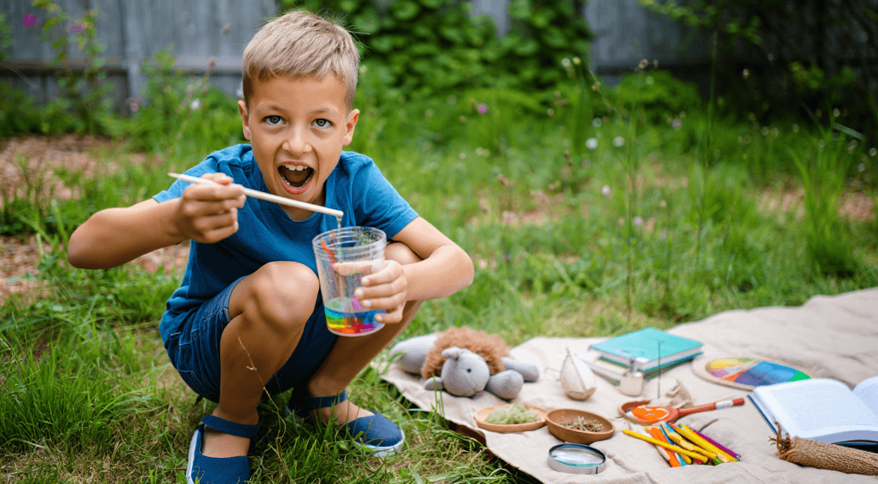 Child playing with rainbow liquid in a cup outdoors, with craft supplies nearby.