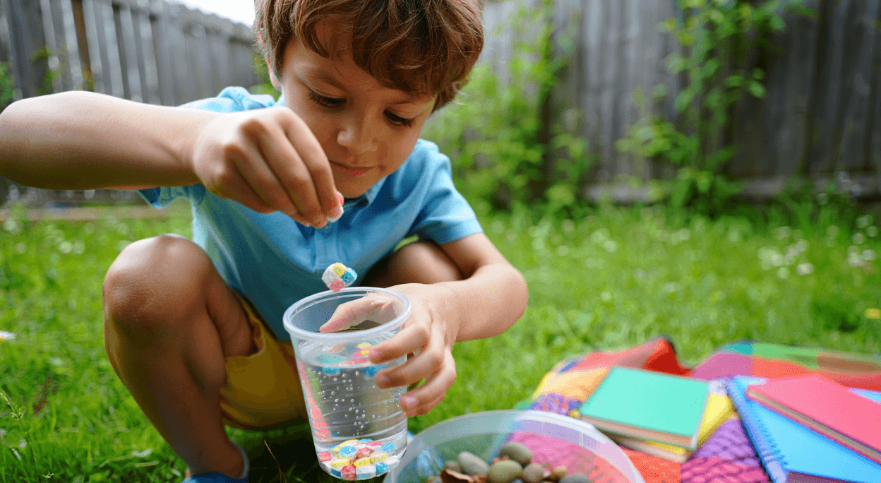 Child dropping colorful squares into a cup of water outdoors.
