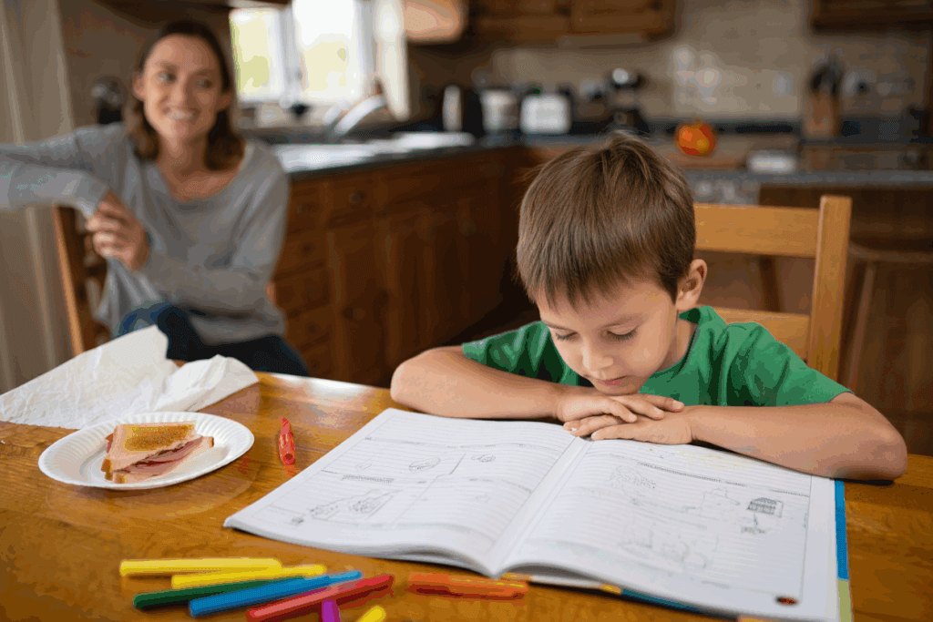 Child studying at a table with an open notebook and an adult in the background.
