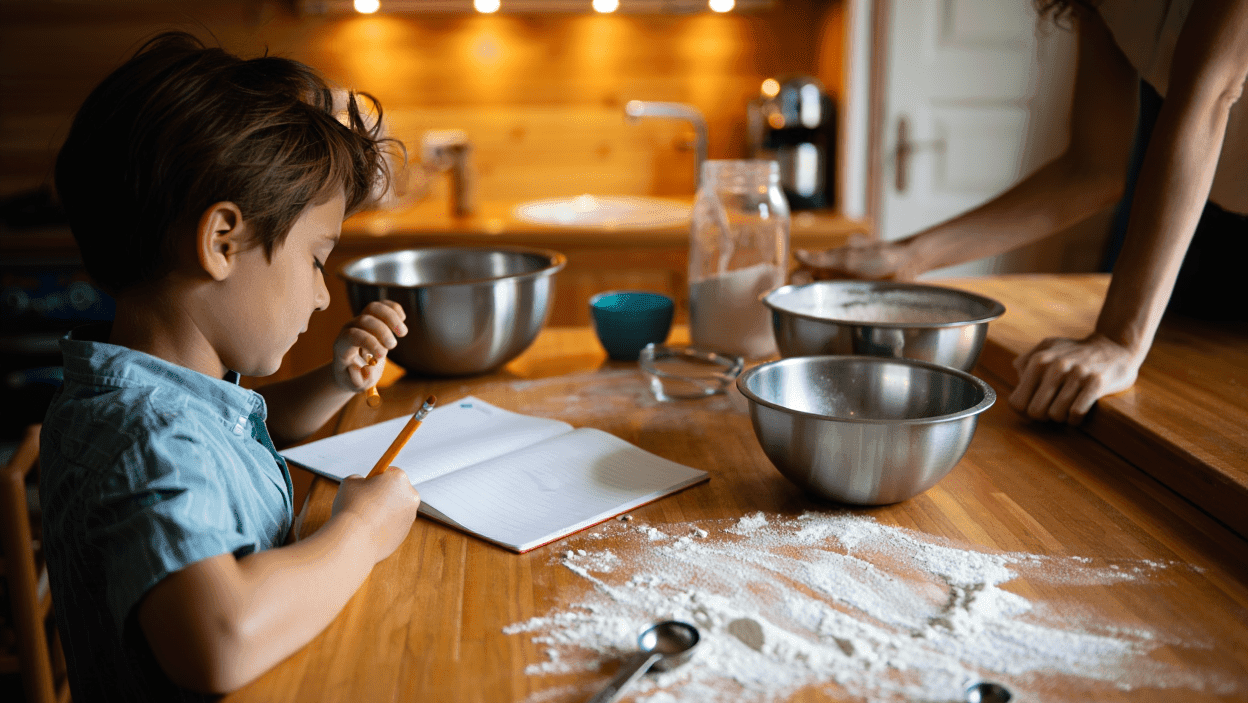 Child writing in a notebook on a flour-dusted kitchen counter.