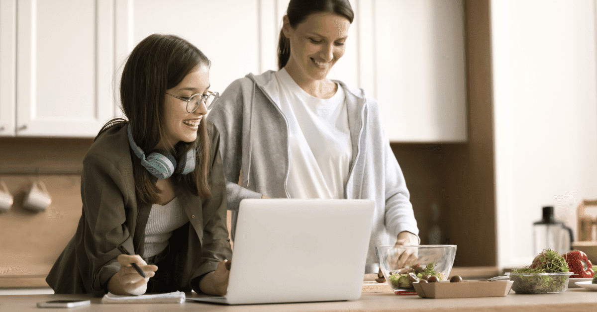 Two adults in a kitchen, one with a laptop, the other preparing food.